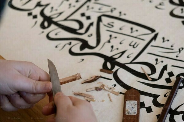 A person's hands, sharpening a wooden tool, with a sheet of Arabic Calligraphy.