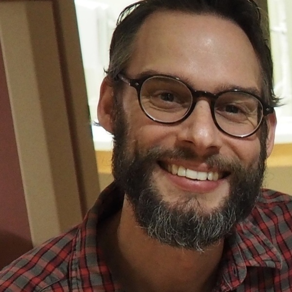 A man with dark hair and a salt-and-pepper beard smiles brightly, showing teeth. He wears dark-rimmed round glasses and a red and grey plaid shirt.